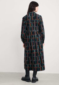 Duga karirana haljina u tamnozelenoj boji s narančastim, bijelim i crvenim akcentima. Ima uski struk, duge rukave i crne čizme do koljena.