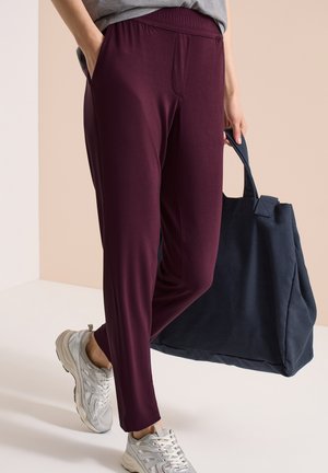 Osoba w bordowych spodniach, szarych sneakersach, niosąca dużą granatową torbę na ramię, stojąca na beżowym tle.