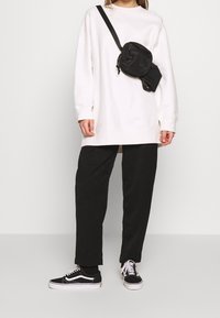 Sudadera blanca de gran tamaño, pantalones negros holgados y zapatillas negras Vans. Se lleva un pequeño bolso negro cruzado sobre el hombro.
