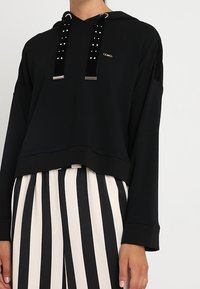 Sudadera cropped de manga larga negra con cordones de terciopelo adornados, combinada con pantalones de tiro alto a rayas verticales en blanco y negro.