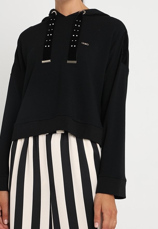 Sudadera cropped de manga larga negra con cordones de terciopelo adornados, combinada con pantalones de tiro alto a rayas verticales en blanco y negro.