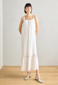 Vestido blanco de algodón con cuerpo fruncido y tirantes ajustables. Presenta un dobladillo con encaje y detalles recortados. Zapatos de tacón bloque plateados.