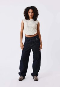 Haut court, beige clair, à coupe décontractée avec un motif texturé et un ourlet arrondi, associé à un jean large bleu foncé avec des coutures blanches.