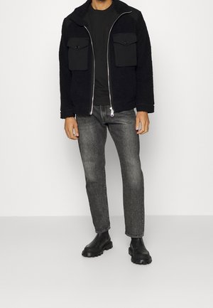 Chaqueta de forro polar negra con dos bolsillos en el pecho, cierre de cremallera y camiseta negra debajo. Se lleva con jeans grises y botas negras.