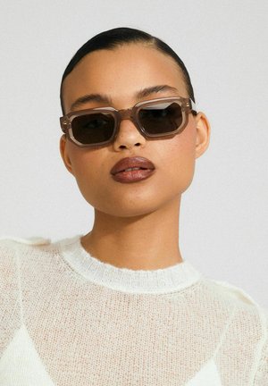 Gafas de sol rectangulares marrones con una montura gruesa y transparente. Las lentes son oscuras, proporcionando un contraste con el suéter claro y con textura que lleva la persona.