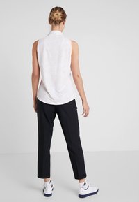 Kvinna med blont hår i en knut, iförd en ärmlös vit mönstrad blus, svarta byxor och vita Adidas-sneakers, stående mot en enfärgad bakgrund.