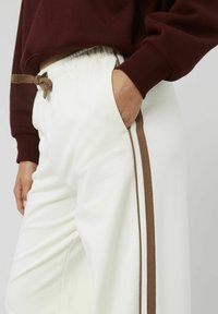 Pantalons de survêtement blancs avec une bande latérale marron, dotés d'une taille élastique et d'un cordon de serrage. Texture lisse avec poches latérales.