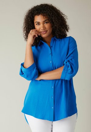 Camicia button-up fresca di colore blu vivace, con maniche arrotolate e un orlo leggermente curvo. Realizzata in un tessuto testurizzato, abbinata a pantaloni bianchi.