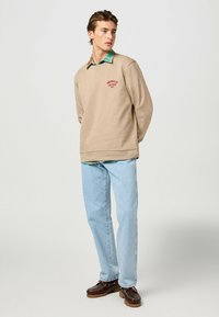 Felpa beige con logo rosso, indossata sopra una camicia a quadri verde e blu. Abbinata a jeans azzurri chiari e scarpe slip-on marroni.