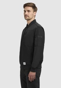 Schwarze Bomberjacke mit Reißverschluss, gerippten cuffs und Saum, Front- und Ärmeltaschen sowie einem strukturierten Stoff. Sichtbares Markenschild an der Unterseite.