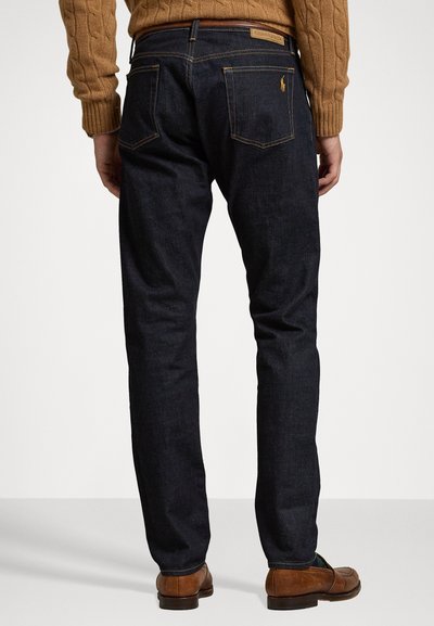 Moški v temno modrih kavbojkah, rjavi pleteni puloverju in rjavih usnjenih loaferjih, stoji s hrbtom obrnjenim proti kameri pred enobarvno ozadjem.