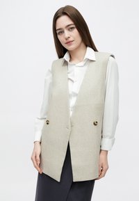 Jeune femme aux longs cheveux bruns portant une chemise blanche boutonnée, un gilet beige sans manches avec des boutons, et une jupe sombre sur un fond blanc.