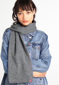 Écharpe en tricot grise drapée autour du cou, fabriquée en tissu doux, associée à une veste en denim bleue. Présente une étiquette en bas.