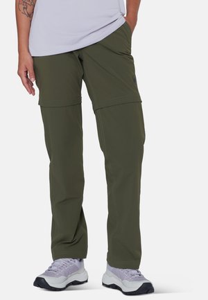 Persona che indossa pantaloni convertibili verde oliva con sezione con zip sulla coscia e sneaker grigio chiaro, in piedi con una mano in tasca.