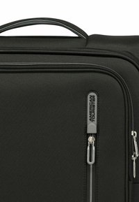 Valigia nera in tessuto con un esterno testurizzato, una maniglia superiore e doppie cerniere con un tirazip brandizzato.