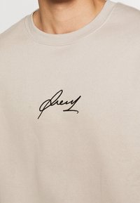 Beige bomulds-sweatshirt med rund halsudskæring og sort broderet signaturlogo på venstre brystområde. Glat tekstur.