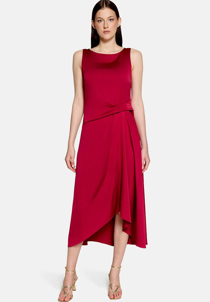 Femme debout portant une robe midi rouge asymétrique sans manches avec un détail drapé et des sandales dorées à talons hauts à brides sur fond blanc.