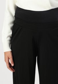 Pantalons noirs avec une ceinture large et pliée, et une texture lisse. Une manche de pull côtelée blanche est visible à côté.