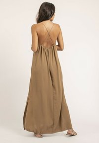 Robe maxi marron sans dos avec une taille froncée, de fines bretelles croisées, des fentes sur les côtés et une texture lisse et fluide.