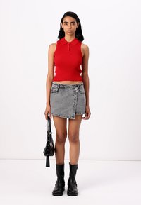 Haut polo rouge à côtes sans manches associé à une mini jupe en denim grise, avec un détail de bouton. Des bottines noires aux chevilles et un sac à bandoulière noir complètent le look.