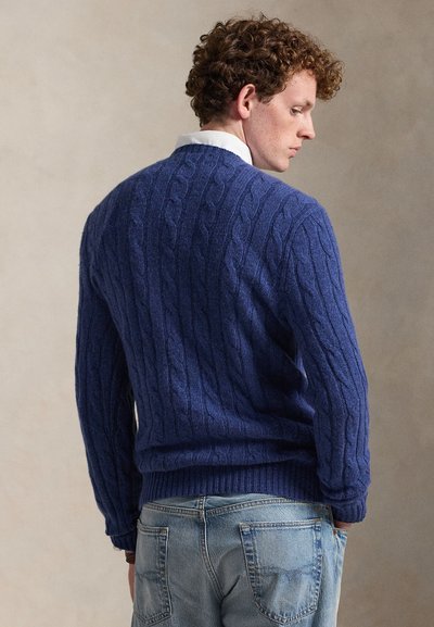 Noore mees lokkis juustega, kandes sinist palmikutega kampsunit valge kraega särgi ja hele siniste teksade kohal, seljaga vaataja poole, vaadates kõrvale.