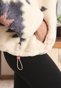 Mano nella tasca di una giacca in pile color crema con dettagli decorati, indossata sopra leggings neri, all'interno vicino a mobili in legno.