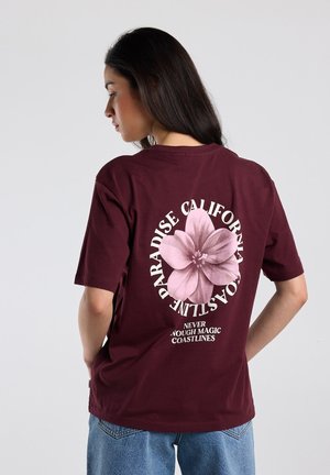 Vrouw met lang donker haar die een bordeauxrood T-shirt draagt met een grote roze bloem en witte tekst over de Californische kustlijnen, gecombineerd met een blauwe spijkerbroek.