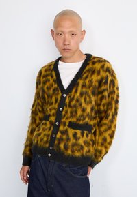 Κάρντιγκ με λεοπάρ σχέδιο σε κίτρινο και μαύρο μείγμα μοχέιρ, με V-λαιμόκοψη, μπροστινές τσέπες και σκούρους κουμπιά. Φορεμένο πάνω από ένα λευκό πουκάμισο.