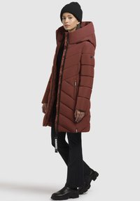 Moteris vilkinti ilgą rudą pūkinę striukę su dideliu apykakle, juodu kepurę, juodas kelnes ir juodus batus, stovinti šonu ant balto fono.