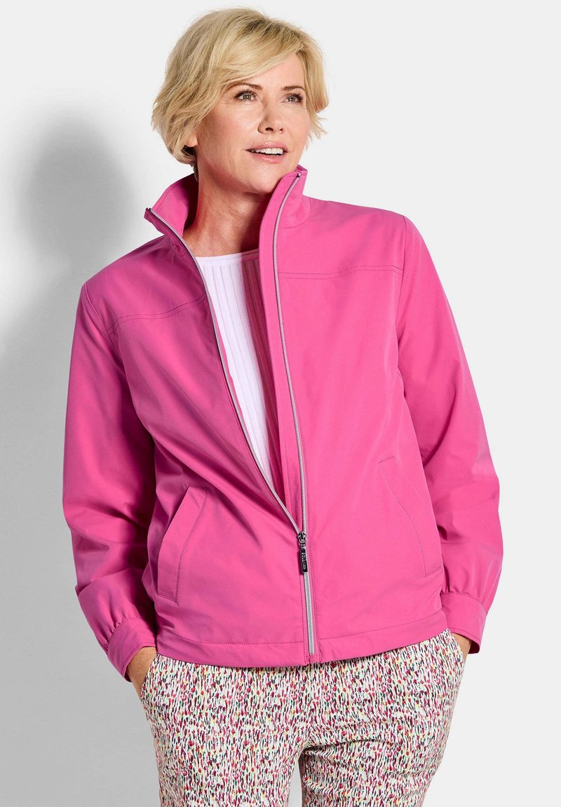 Femme aux cheveux blonds courts portant une veste zippée rose vif et un pantalon à motifs, debout les mains dans les poches, regardant légèrement vers le haut et à droite.