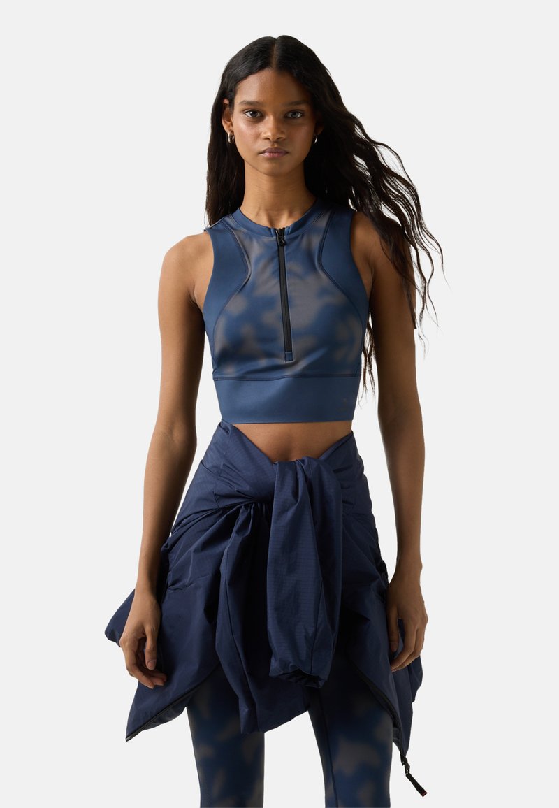 Kvinde med langt hår iført navyblåt ærmeløst croptop med lynlås foran, bundet navyblå jakke omkring livet og matchende leggings.