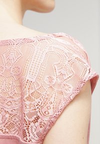 Růžový top s jemnými krajkovými detaily na ramenou, který ukazuje složité vzory. Měkká, hladká látka s uvolněným střihem a krátkými rukávy.