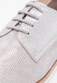 Chaussure à lacets en argent métallique clair avec une texture perforée, des lacets gris et une semelle marron. Dotée d'une tige en cuir lisse et d'un design classique.