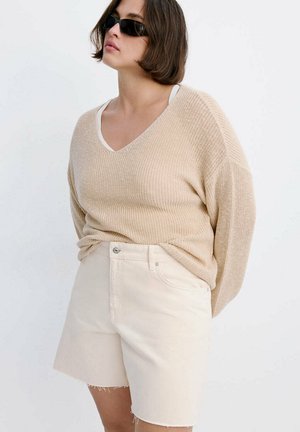 Femeie care poartă un pulover tricotat bej și pantaloni scurți off-white cu tiv nefinisat, stând pe un fundal alb simplu, purtând ochelari de soare negri.