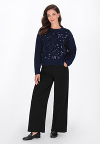 Vrouw staat met één hand in de zak, draagt een marineblauwe pailletten trui, zwarte wijde broek en zwarte instapschoenen op een witte achtergrond.