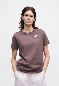 Γυναίκα με καστανά μαλλιά μέχρι τους ώμους στέκεται με τα χέρια στις τσέπες, φορώντας ένα μοβ t-shirt της Nike και ανοιχτό ροζ σορτσάκι, με φόντο λευκό.