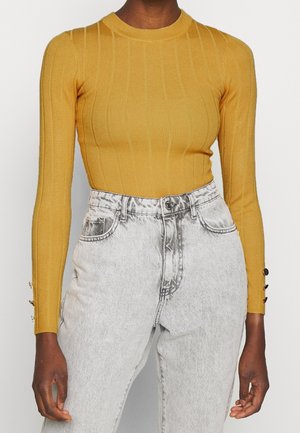 Personne portant un haut à manches longues côtelé jaune moutarde avec des boutons sur les manches et un jean taille haute gris clair délavé à l'acide.