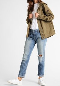 Olivgrön, huoodad jacka med tryckknappar och dragkedja. Buren över en vit skjorta, tillsammans med ljusblå ripped jeans och vita sneakers.