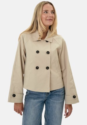 Femme portant une veste beige croisée avec des boutons noirs et des manches larges, associée à un jean bleu, regardant légèrement sur le côté.