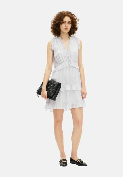 Jonge vrouw in een mouwloze witte jurk met franje, die een zwarte clutch met studs vasthoudt en zwarte puntige ballerina's draagt.