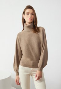 Beige kõrge kaelusega kootud särk, millel on paelavad varrukad, koos kreemikatest pükstest kerge tausta ees.