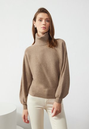 Pull à col roulé beige en maille côtelée avec des manches ballons, associé à un pantalon couleur crème sur un fond clair.