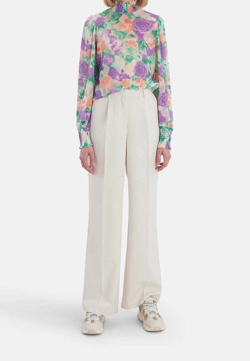 Blouse floral à manches longues en violet, orange et vert, associée à un pantalon large blanc et des baskets beiges. Tissu lisse avec des accents froncés.