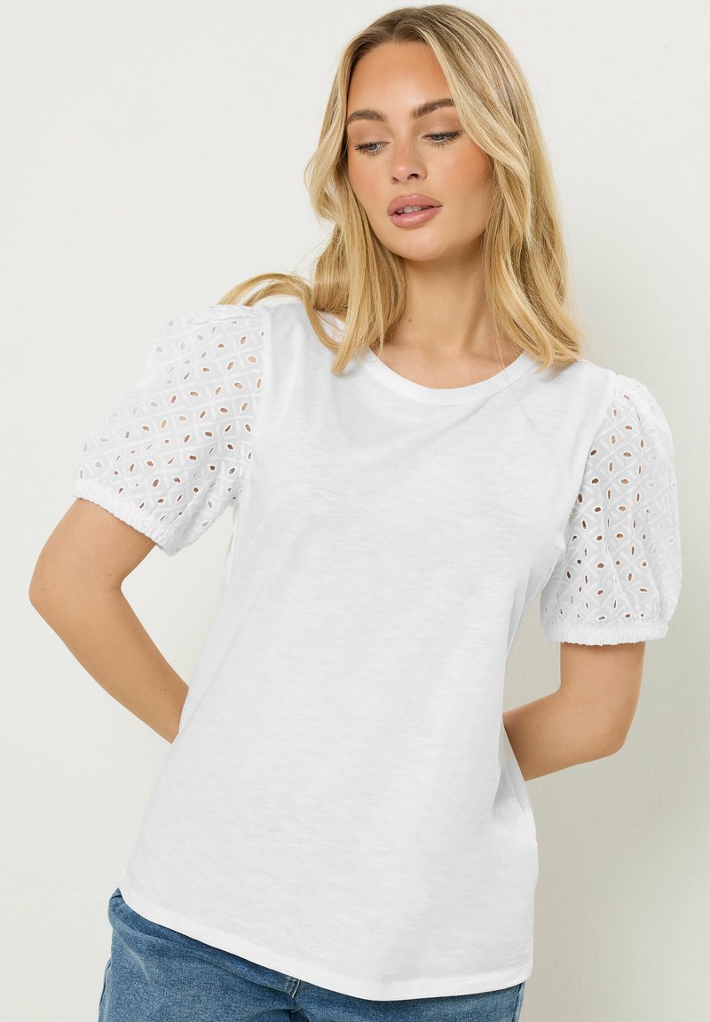 Femme aux cheveux blonds portant un t-shirt blanc à manches bouffantes ajourées, debout avec une main sur la hanche devant un fond uni.