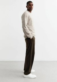 Pull en maille beige texturé avec un col rond, associé à un pantalon ample vert olive et des baskets blanches. Le modèle se tient dans un cadre neutre.