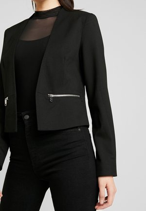 Frau trägt einen taillierten schwarzen Crop-Blazer mit silbernen Reißverschlusstaschen über einem schwarzen transparenten Top und schwarzen hoch taillierten Hosen.