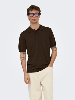 Jeune homme tatoué portant un polo à manches courtes marron foncé et un pantalon couleur crème, debout avec une main dans la poche.