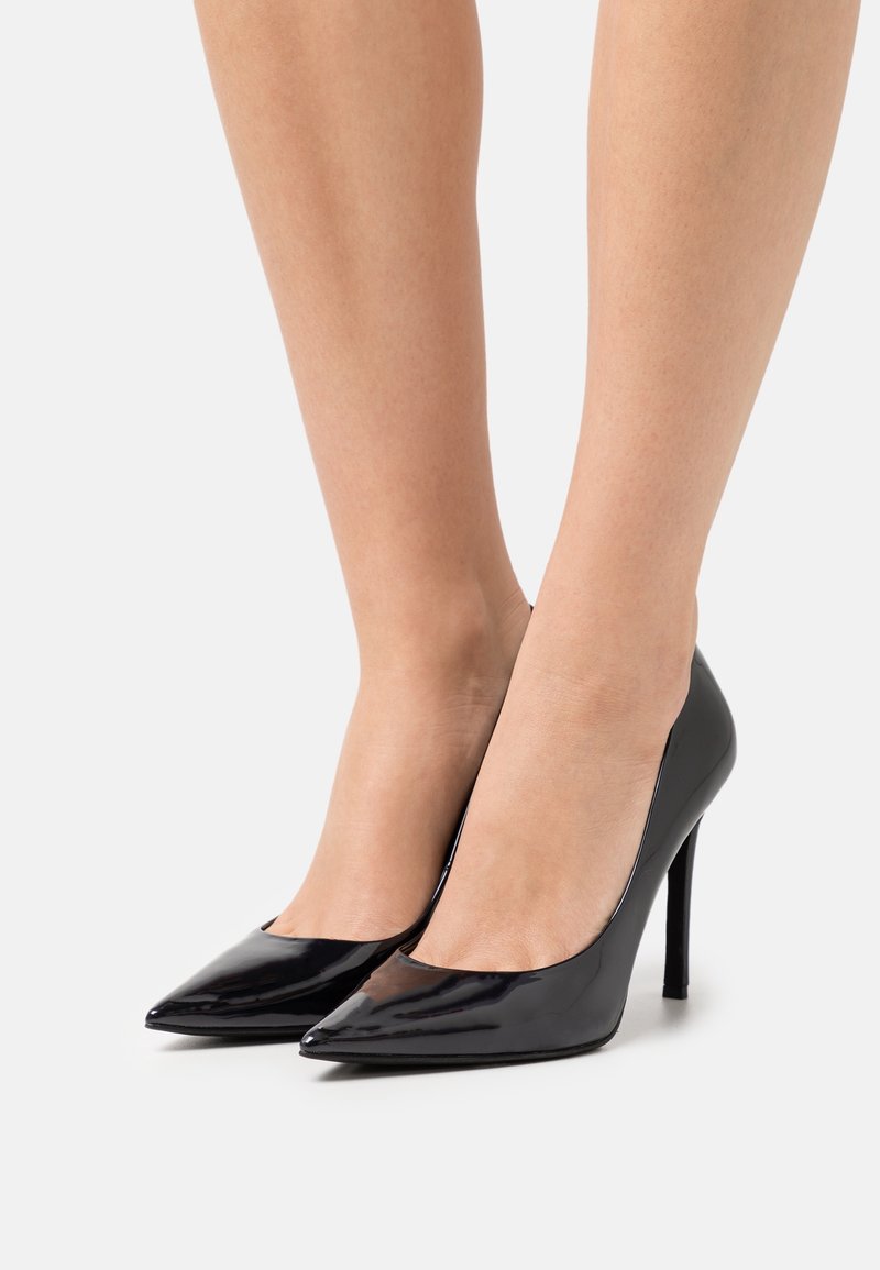 Paire d'escarpins pointus noirs brillants à talons aiguilles portés pieds nus sur un fond clair uni.
