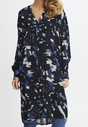 Femme portant une robe tunique à manches longues boutonnée avec un motif abstrait noir, bleu, gris et blanc, associée à un jean bleu.