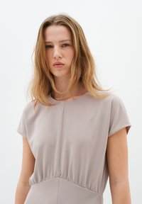 Šaty s krátkým rukávem ve světle taupe látce. Mají kulatý výstřih, nařasený pas a hladkou texturu. Jednoduchý, minimalistický design.